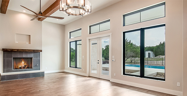 Modern custom home living room with floor-to-ceiling windows, exposed wood beams, fireplace, and pool views in Boerne, Texas